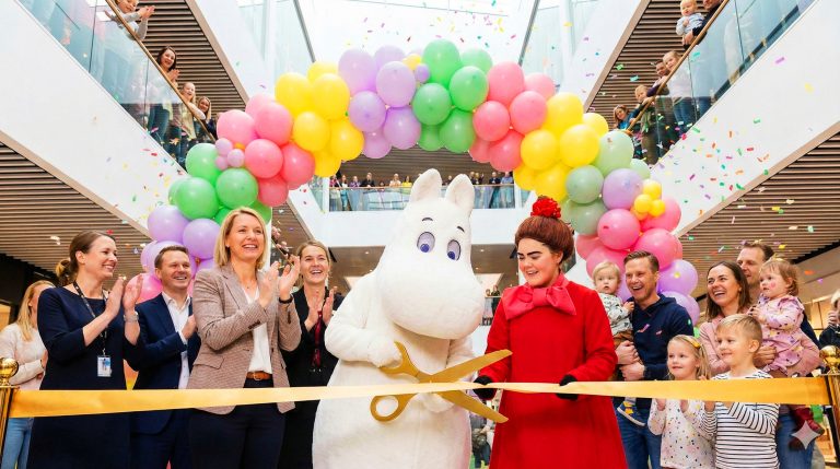 Muumipeikko ja Pikku Myy leikkaavat avajaisnauhaa isoilla saksilla. Taustalla yleisöä ja ilmapalloja.