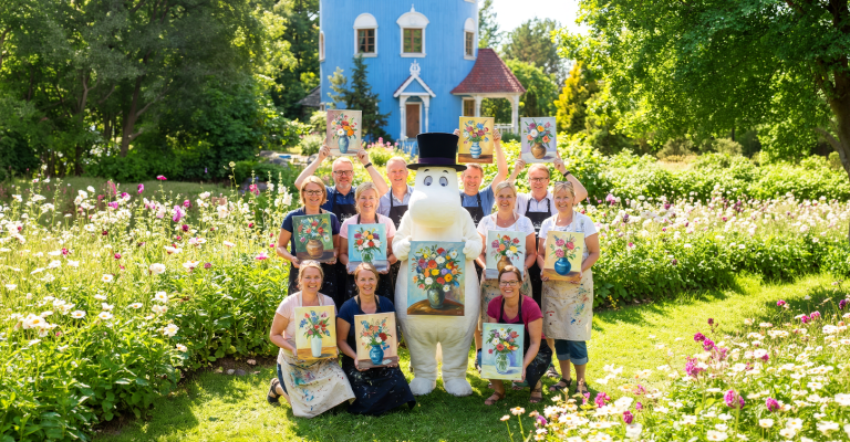 Muumipappa ja ryhmä aikuisia Muumitaon edustalla maalaukset käsissä.